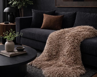 Könnte beinhalten: Ein gemütliches Wohnzimmer mit einem dunkelgrauen Sofa, schwarzen und braunen Kissen und einer hellbraunen, flauschigen Decke. Ein schwarzer Couchtisch und ein Teppich ergänzen das Arrangement. Dunkle, stimmungsvolle Atmosphäre.