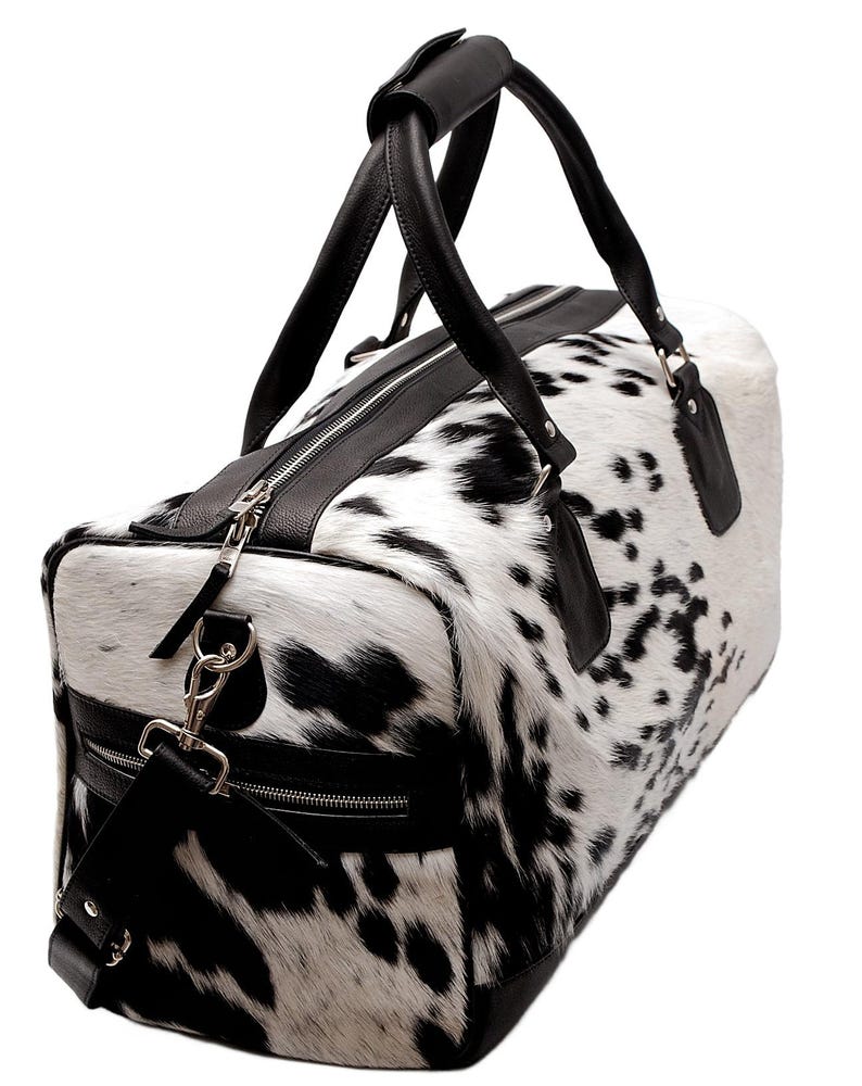 K&ouml;nnte beinhalten: Eine Reisetasche mit Kuhfellmuster in Schwarz und Wei&szlig;. Die Tasche hat schwarze Ledergriffe, Zierleisten und einen Schultergurt. Die Tasche hat einen Rei&szlig;verschluss und Metallbeschl&auml;ge.