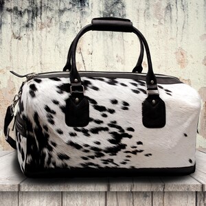 K&ouml;nnte beinhalten: Eine schwarz-wei&szlig; gefleckte Reisetasche mit schwarzen Ledergriffen und -besatz. Die Tasche hat einen Rei&szlig;verschluss und einen abnehmbaren Schultergurt. Die Tasche ist aus Kuhfell.