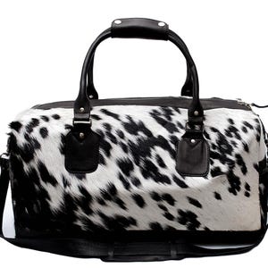 K&ouml;nnte beinhalten: Eine Reisetasche mit Kuhfellmuster in Schwarz und Wei&szlig;. Die Tasche hat schwarze Ledergriffe und einen Schultergurt. Die Tasche ist f&uuml;r Reisen konzipiert und hat einen Rei&szlig;verschluss. Die Abmessungen sind nicht sichtbar.
