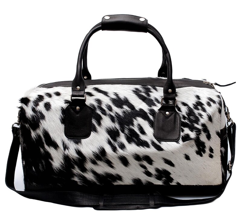 K&ouml;nnte beinhalten: Eine Reisetasche mit Kuhfellmuster in Schwarz und Wei&szlig;. Die Tasche hat schwarze Ledergriffe und einen Schultergurt. Die Tasche ist f&uuml;r Reisen konzipiert und hat einen Rei&szlig;verschluss. Die Abmessungen sind nicht sichtbar.