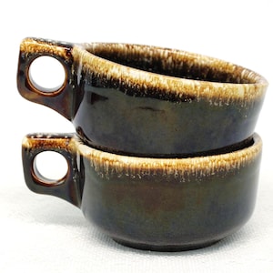 May include: Two stacked, dark brown ceramic soup bowls with a handle and a circular opening. The rims have a lighter brown, textured glaze. The bowls are on a white surface.