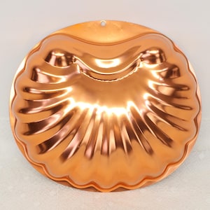 May include: A copper-colored, round bundt pan with a scalloped edge and a seashell design. The pan has a shiny finish and a small hole at the top for hanging. The baking pan is designed for making cakes and other baked goods.