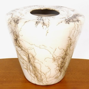 May include: A white vase with a unique design. The vase has a wide, rounded top and tapers down to a narrower base. The surface is decorated with a network of black, vein-like lines. The vase is sitting on a wooden surface.