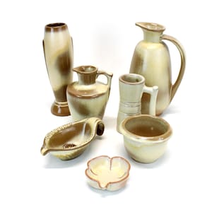 May include: A collection of ceramic pottery in shades of cream and brown. The assortment includes a tall vase, a pitcher with a handle, a small pitcher, a mug, a small bowl, a leaf-shaped dish, and a flower-shaped dish.