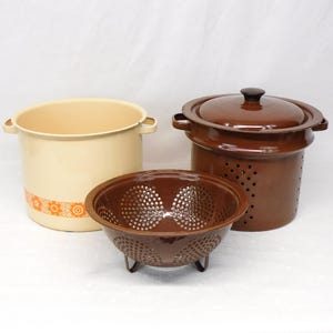 May include: A collection of vintage kitchenware, including a cream-colored enamel pot with orange floral accents, a brown colander, and a brown enamel pot with a lid. The items are arranged on a white surface.