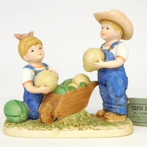May include: A ceramic figurine depicting two children in denim overalls and straw hats, carrying melons. One child pushes a wooden wheelbarrow filled with melons. The base is painted to resemble grass. The figurine is labeled "DENIM DAYS".