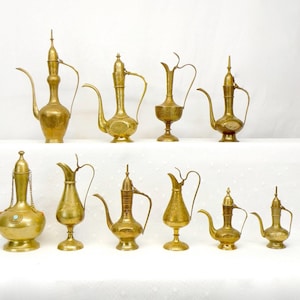 May include: A collection of antique brass pitchers and ewers, each with a unique design. The pitchers vary in size and shape, featuring spouts, handles, and decorative detailing. Some have ornate lids and bases, showcasing a range of styles.