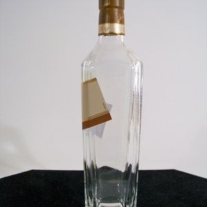 May include: A clear glass bottle with a brown cap and a gold label. The bottle is empty and has a rectangular shape.