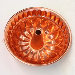 May include: A copper-colored Bundt cake pan with a central tube and decorative geometric and floral patterns. The pan has a shiny finish and a small handle on the side. This baking pan is designed for making cakes.