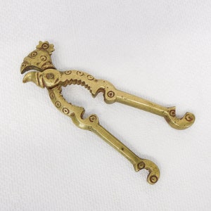 May include: A vintage brass nutcracker with a bird-like head and decorative detailing. The handles are curved with circular accents. The nutcracker is open, ready for use, and sits on a white surface.