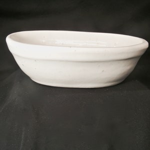 May include: A white, oval-shaped ceramic bowl. The bowl has a simple, classic design with a slightly raised rim. The smooth surface of the bowl reflects the light, highlighting its clean lines. The bowl is suitable for serving food.
