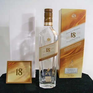May include: A clear glass bottle of Johnnie Walker 18 year old blended Scotch whisky. The bottle has a gold label with the Johnnie Walker logo and the words "Aged 18 Years". The bottle is sitting on a black surface next to a gold box with the same label.
