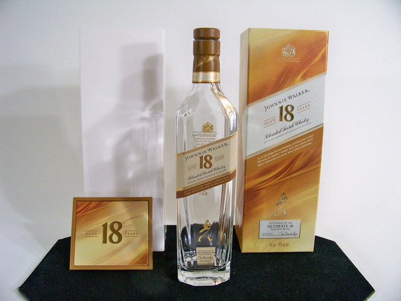 May include: A clear glass bottle of Johnnie Walker 18 year old blended Scotch whisky. The bottle has a gold label with the Johnnie Walker logo and the words "Aged 18 Years". The bottle is sitting on a black surface next to a gold box with the same label.