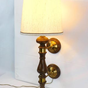 May include: A pair of vintage wall sconces with a wooden base and brass accents. Each sconce features a cream-colored lampshade with a brown trim. The sconces are designed to be mounted on a wall and have a classic, elegant style.