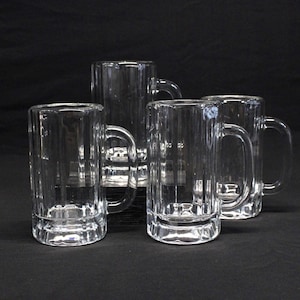 May include: Four clear glass beer mugs with vertical ribbed sides and sturdy handles are arranged against a black background. The mugs are empty and appear to be made of thick glass, suitable for serving cold beverages.