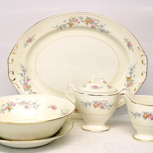 May include: A cream-colored porcelain dinnerware set featuring a large oval platter, a serving bowl with a saucer, a covered sugar bowl, and a creamer. Each piece is adorned with a delicate floral pattern in pink, yellow, and blue, with gold trim.