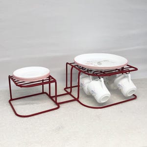 May include: A red metal serving stand with two tiers. The top tier holds a stack of white plates with pink rims. The lower tier has hooks for hanging white teacups with floral designs. The stand is designed to hold a tea set.