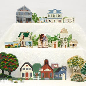 May include: A collection of hand-painted wooden miniature houses and buildings, including a general store, a schoolhouse, a church, and a few homes. The houses are arranged in a row on a white surface, with trees and other details surrounding them.
