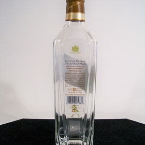 May include: A clear glass bottle of Johnnie Walker Blended Scotch Whisky. The bottle has a gold label with the Johnnie Walker logo and text. The label also includes the government warning about alcohol consumption during pregnancy. The bottle is sitting on a white surface.