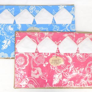 May include: Two packages of table linens. One is blue with white floral patterns, the other is pink with white floral patterns. Each package contains white napkins. The packages are rectangular with a gold border.