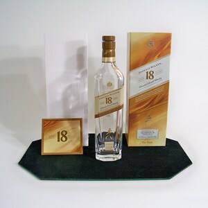 May include: A clear glass bottle of Johnnie Walker 18 year old blended Scotch whisky with a gold label. The bottle is sitting on a black tray with a gold card that says "18 years" on it. The bottle is next to a gold box with the Johnnie Walker logo and the text "18 years old blended Scotch whisky" on it.
