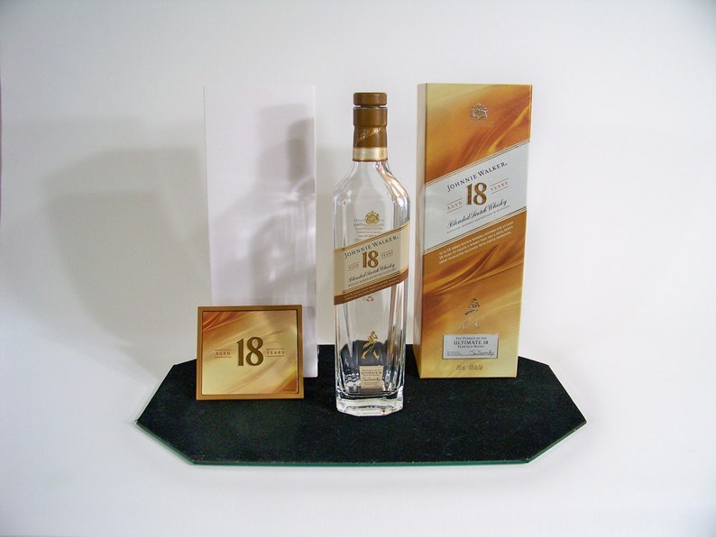 May include: A clear glass bottle of Johnnie Walker 18 year old blended Scotch whisky with a gold label. The bottle is sitting on a black tray with a gold card that says "18 years" on it. The bottle is next to a gold box with the Johnnie Walker logo and the text "18 years old blended Scotch whisky" on it.