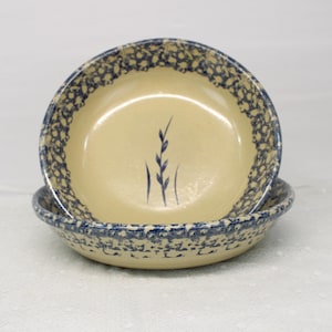 May include: Two ceramic bowls with a cream-colored interior and a blue speckled rim. The bowls are stacked, with the top bowl featuring a blue wheat stalk design. The bowls are suitable for serving soups, salads, or desserts.