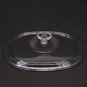 May include: Clear glass oval-shaped lid with a small, round handle. The lid has a decorative, ridged pattern in the center and a smooth, rounded edge. The lid is designed for cookware.