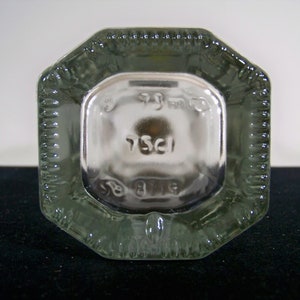 May include: Close-up of the bottom of a clear glass bottle with an octagonal shape. The bottom of the bottle has a raised, textured design and the inscription "75cl" is visible.