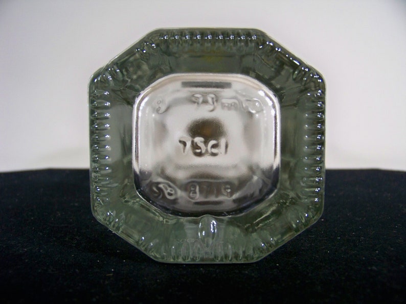 May include: Close-up of the bottom of a clear glass bottle with an octagonal shape. The bottom of the bottle has a raised, textured design and the inscription "75cl" is visible.