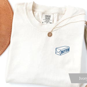 May include: Ivory-colored t-shirt with a blue and white butter graphic. The shirt has a crew neck and short sleeves. A gold necklace with a pendant is draped across the shirt. A brown hat and dried plant are in the background.