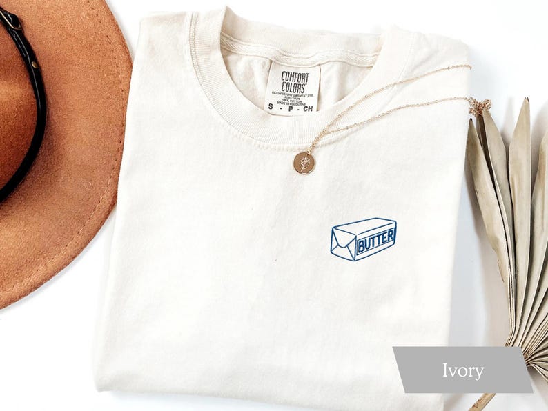 May include: Ivory-colored t-shirt with a blue and white butter graphic. The shirt has a crew neck and short sleeves. A gold necklace with a pendant is draped across the shirt. A brown hat and dried plant are in the background.