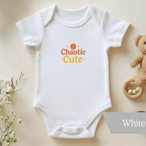 May include: White baby onesie with the text "Chaotic Cute" in orange and yellow. A small teddy bear and a wooden teething ring with white beads are next to the onesie. The onesie is a short-sleeved bodysuit.