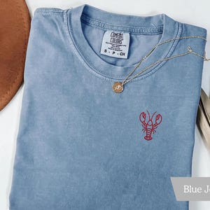K&ouml;nnte beinhalten: Ein blaues Jeans-T-Shirt mit einem roten Hummer, der auf der Vorderseite gestickt ist. Das Shirt hat einen Rundhalsausschnitt und ein kleines Etikett am Kragen. Eine goldene Halskette mit Anh&auml;nger liegt &uuml;ber dem Shirt.