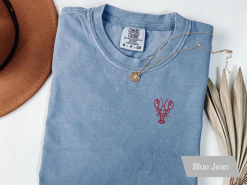 K&ouml;nnte beinhalten: Ein blaues Jeans-T-Shirt mit einem roten Hummer, der auf der Vorderseite gestickt ist. Das Shirt hat einen Rundhalsausschnitt und ein kleines Etikett am Kragen. Eine goldene Halskette mit Anh&auml;nger liegt &uuml;ber dem Shirt.