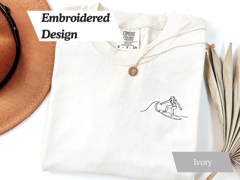 May include: Ivory-colored t-shirt with an embroidered black line drawing of a surfer riding a wave. The shirt has the words "Embroidered Design" in a white banner. A gold necklace is draped on the shirt.