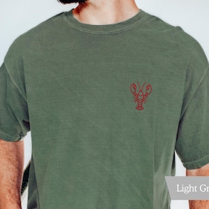 K&ouml;nnte beinhalten: Hellgr&uuml;nes T-Shirt mit einer kleinen roten Hummerstickerei auf der Brust. Das Shirt hat kurze &Auml;rmel und einen Rundhalsausschnitt. Der Stoff wirkt weich und leicht verwaschen.