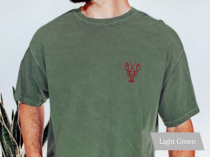 K&ouml;nnte beinhalten: Hellgr&uuml;nes T-Shirt mit einer kleinen roten Hummerstickerei auf der Brust. Das Shirt hat kurze &Auml;rmel und einen Rundhalsausschnitt. Der Stoff wirkt weich und leicht verwaschen.