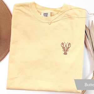 K&ouml;nnte beinhalten: Hellgelbes T-Shirt mit roter gestickter Hummer-Design. Das Shirt hat einen Rundhalsausschnitt und kurze &Auml;rmel. Der Text "COMFORT FINDS" ist auf dem Etikett sichtbar. Eine goldene Halskette mit Sternanh&auml;nger liegt auf dem Shirt. Das Wort "Butter" steht unten rechts.