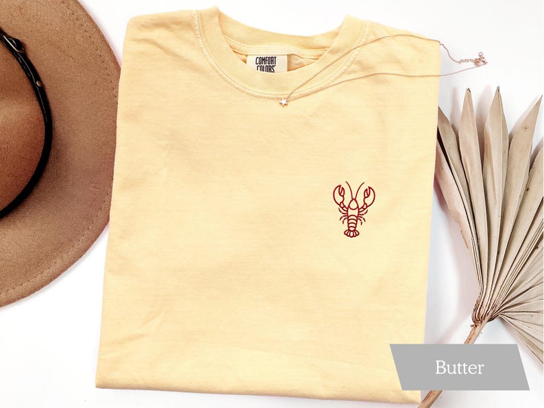 K&ouml;nnte beinhalten: Hellgelbes T-Shirt mit roter gestickter Hummer-Design. Das Shirt hat einen Rundhalsausschnitt und kurze &Auml;rmel. Der Text "COMFORT FINDS" ist auf dem Etikett sichtbar. Eine goldene Halskette mit Sternanh&auml;nger liegt auf dem Shirt. Das Wort "Butter" steht unten rechts.
