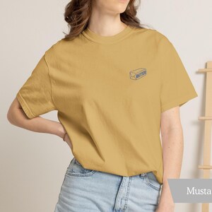 May include: Mustard-colored t-shirt with a small blue illustration of a butter stick on the chest. The shirt is made of soft material and is paired with light blue denim shorts. The word "Mustard" is displayed on a gray banner.
