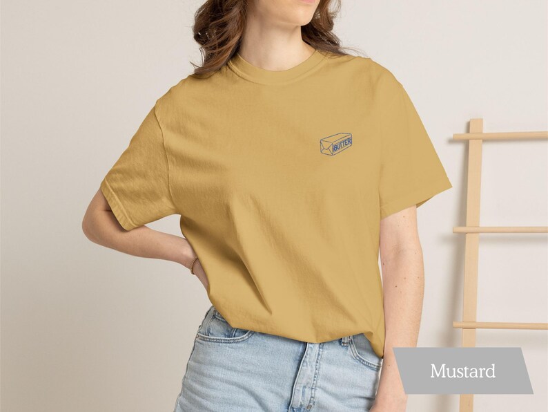 May include: Mustard-colored t-shirt with a small blue illustration of a butter stick on the chest. The shirt is made of soft material and is paired with light blue denim shorts. The word "Mustard" is displayed on a gray banner.