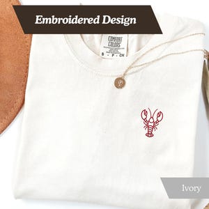 K&ouml;nnte beinhalten: Elfenbeinfarbenes T-Shirt mit rotem, gesticktem Hummer-Design. Das Shirt hat einen Rundhalsausschnitt und kurze &Auml;rmel. Die Worte "Embroidered Design" werden oben links auf dem Shirt angezeigt.