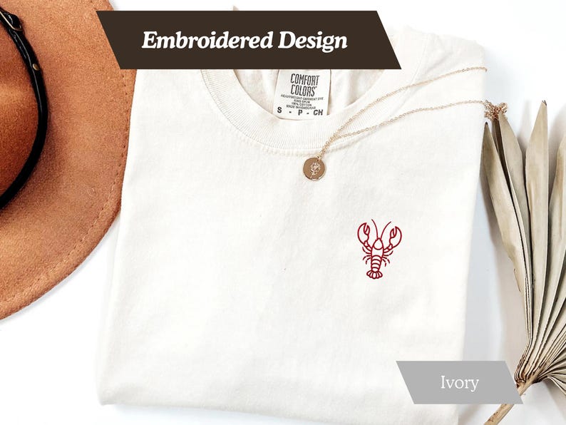 K&ouml;nnte beinhalten: Elfenbeinfarbenes T-Shirt mit rotem, gesticktem Hummer-Design. Das Shirt hat einen Rundhalsausschnitt und kurze &Auml;rmel. Die Worte "Embroidered Design" werden oben links auf dem Shirt angezeigt.