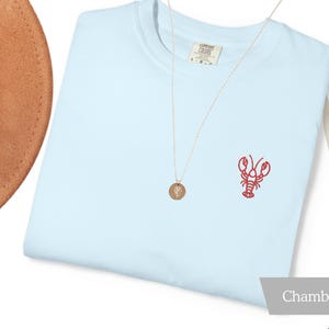 K&ouml;nnte beinhalten: Hellblaues T-Shirt mit rotem, gesticktem Hummer-Design auf der Vorderseite. Eine goldene Halskette mit Anh&auml;nger und ein brauner Hut sind ebenfalls zu sehen. Das Shirt ist mit "Chambray" beschriftet.
