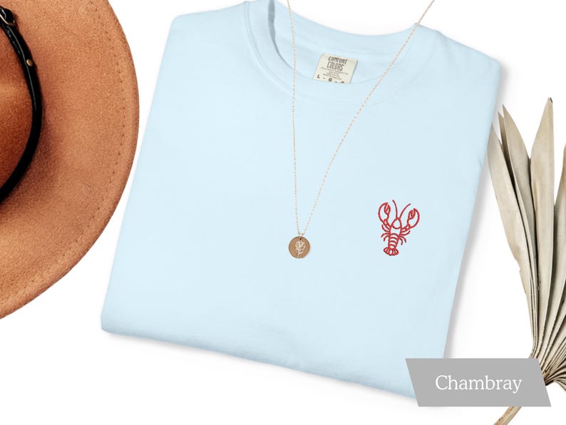 K&ouml;nnte beinhalten: Hellblaues T-Shirt mit rotem, gesticktem Hummer-Design auf der Vorderseite. Eine goldene Halskette mit Anh&auml;nger und ein brauner Hut sind ebenfalls zu sehen. Das Shirt ist mit "Chambray" beschriftet.