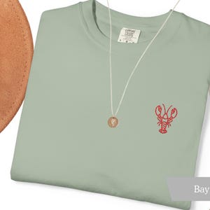 K&ouml;nnte beinhalten: Hellgr&uuml;nes T-Shirt mit einer roten Hummerstickerei auf der Brust. Eine goldene Anh&auml;ngerkette und ein brauner Hut sind ebenfalls zu sehen. Das T-Shirt ist mit "Comfort Colors" gekennzeichnet. Ein dekoratives Palmblatt befindet sich im Hintergrund.