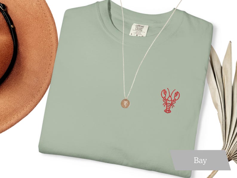 K&ouml;nnte beinhalten: Hellgr&uuml;nes T-Shirt mit einer roten Hummerstickerei auf der Brust. Eine goldene Anh&auml;ngerkette und ein brauner Hut sind ebenfalls zu sehen. Das T-Shirt ist mit "Comfort Colors" gekennzeichnet. Ein dekoratives Palmblatt befindet sich im Hintergrund.