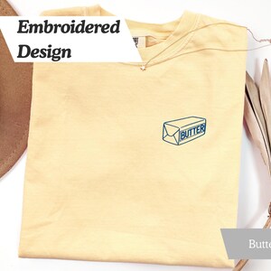 May include: A light yellow t-shirt with an embroidered butter design. The design is a blue outline of a butter package with the word "Butter" inside. The shirt also has the words "Embroidered Design" in a white banner.
